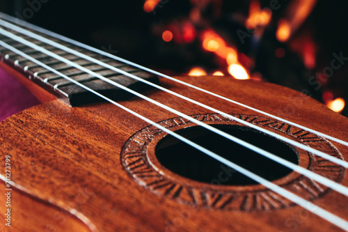 Ukulele small guitar close up stings, fireplace on the background. Musical concept, guitar fret board macro, fire in chimney, cosy romantic atmosphere.