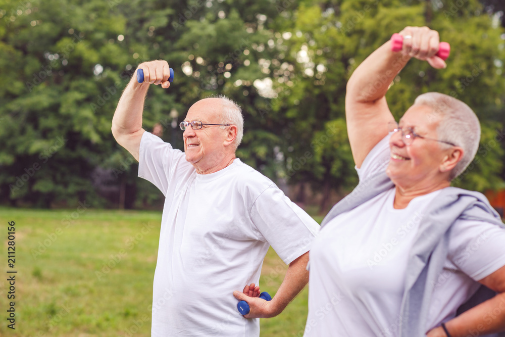 senior man and woman are outdoors in a park exercise with dumbbells and having fun together.