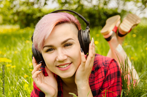 Young beautiful girl listening to music on headphones and lies on a green grass and dandelions.