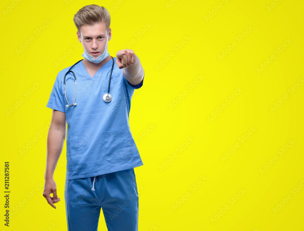 Young handsome blond doctor pointing with finger to the camera and to you, hand sign, positive and confident gesture from the front Young handsome blond doctor pointing with finger to the camera and to you, hand sign, positive and confident gesture from the front