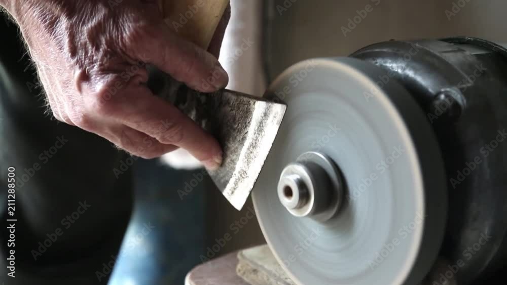 Hands hold an ax near the abrasive machine. The man sharpens the ax blade on the machine. Hands hold an ax near the abrasive machine. The man sharpens the ax blade on the machine.