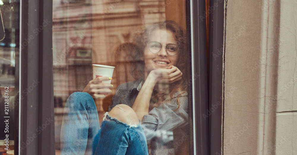 Young redhead curly girl wearing casual clothes and glasses sitting on a window sill with a takeaway coffee. Young redhead curly girl wearing casual clothes and glasses sitting on a window sill with a takeaway coffee.