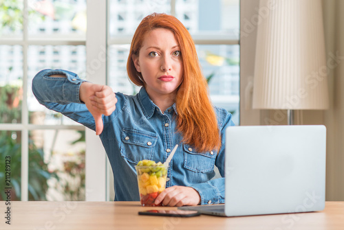 Redhead woman using computer laptop eating fruit at home with angry face, negative sign showing dislike with thumbs down, rejection concept