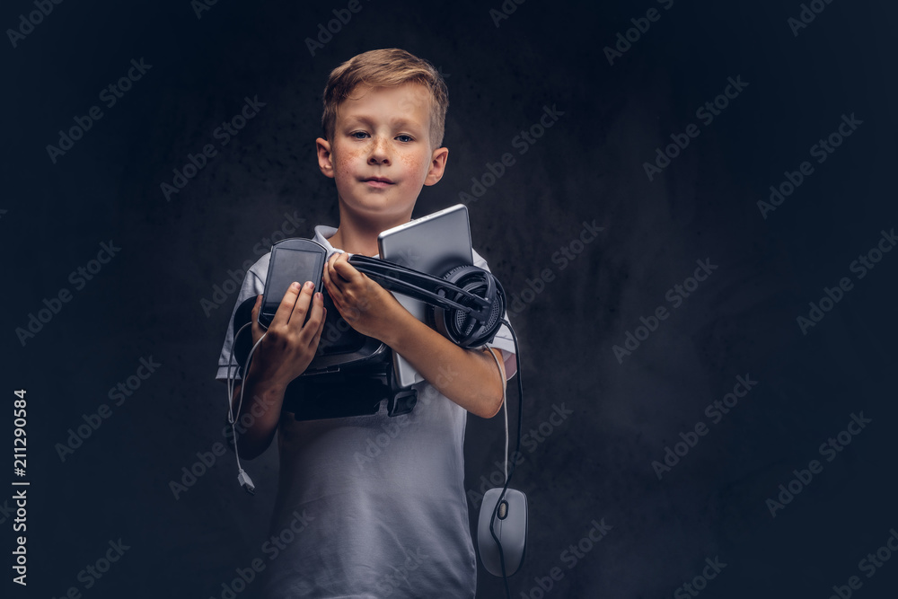 Cute schoolboy dressed in a white t-shirt, holds full digital set for entertainment at a studio. Isolated on dark textured background.