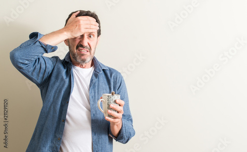 Senior man drinking coffee in a mug stressed with hand on head, shocked with shame and surprise face, angry and frustrated. Fear and upset for mistake.