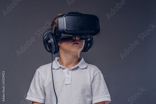 Close-up portrait of a schoolboy dressed in a white t-shirt standing in a virtual reality glasses at a studio. Isolated on gray background.