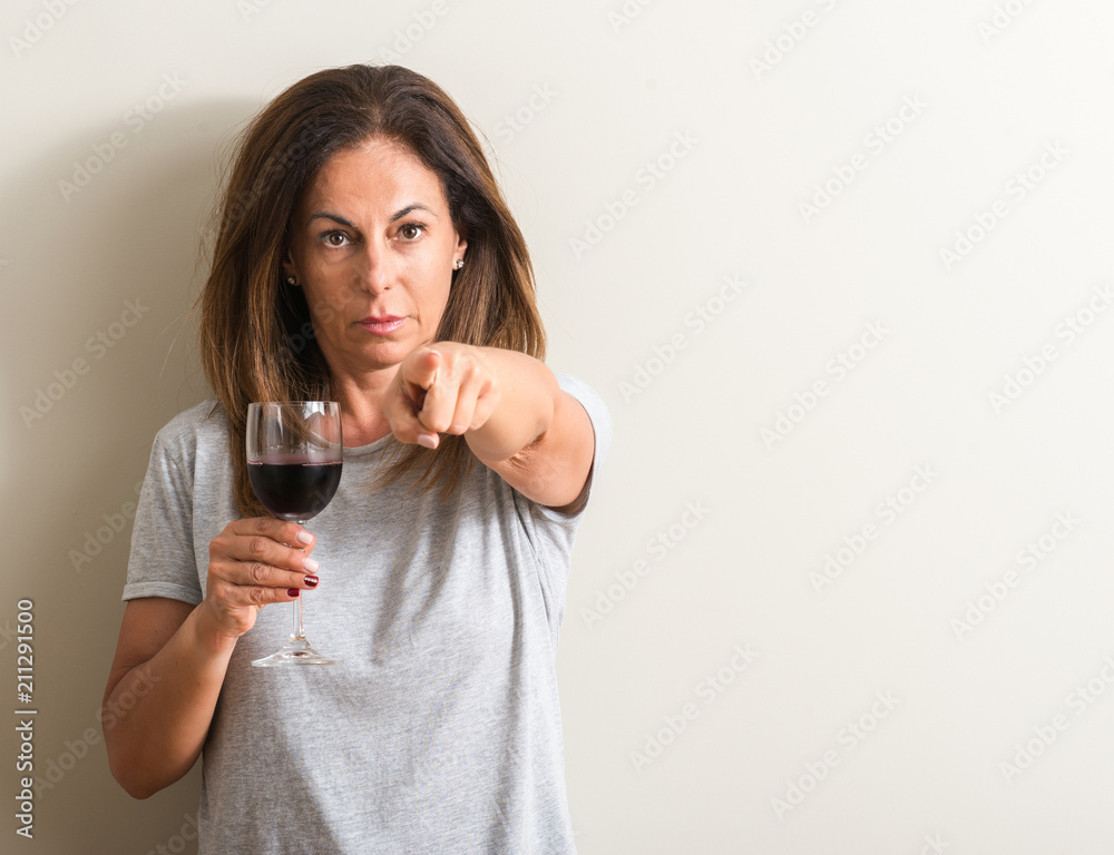 Middle age woman drinking red wine in a glass pointing with finger to the camera and to you, hand sign, positive and confident gesture from the front Middle age woman drinking red wine in a glass pointing with finger to the camera and to you, hand sign, positive and confident gesture from the front