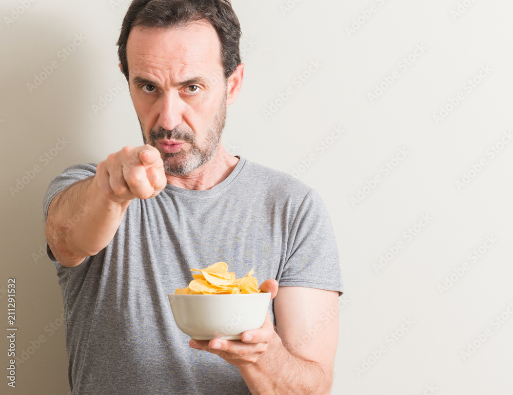 Senior man eating potato chips pointing with finger to the camera and to you, hand sign, positive and confident gesture from the front Senior man eating potato chips pointing with finger to the camera and to you, hand sign, positive and confident gesture from the front