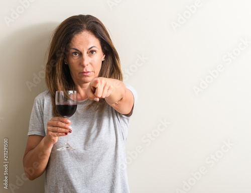 Middle age woman drinking red wine in a glass pointing with finger to the camera and to you, hand sign, positive and confident gesture from the front