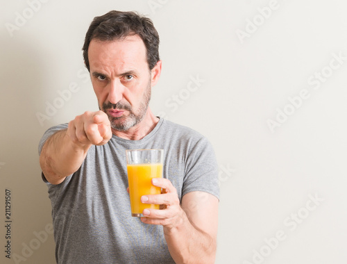 Senior man drinking orange juice in a glass pointing with finger to the camera and to you, hand sign, positive and confident gesture from the front