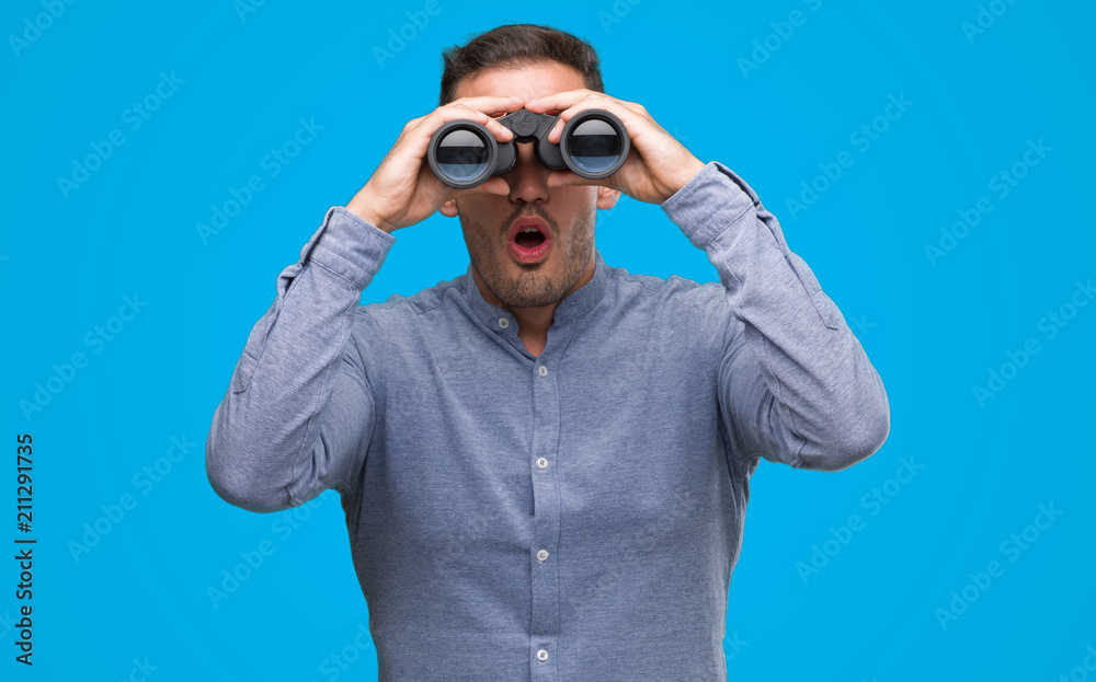 Handsome young man looking through binoculars scared in shock with a surprise face, afraid and excited with fear expression Handsome young man looking through binoculars scared in shock with a surprise face, afraid and excited with fear expression