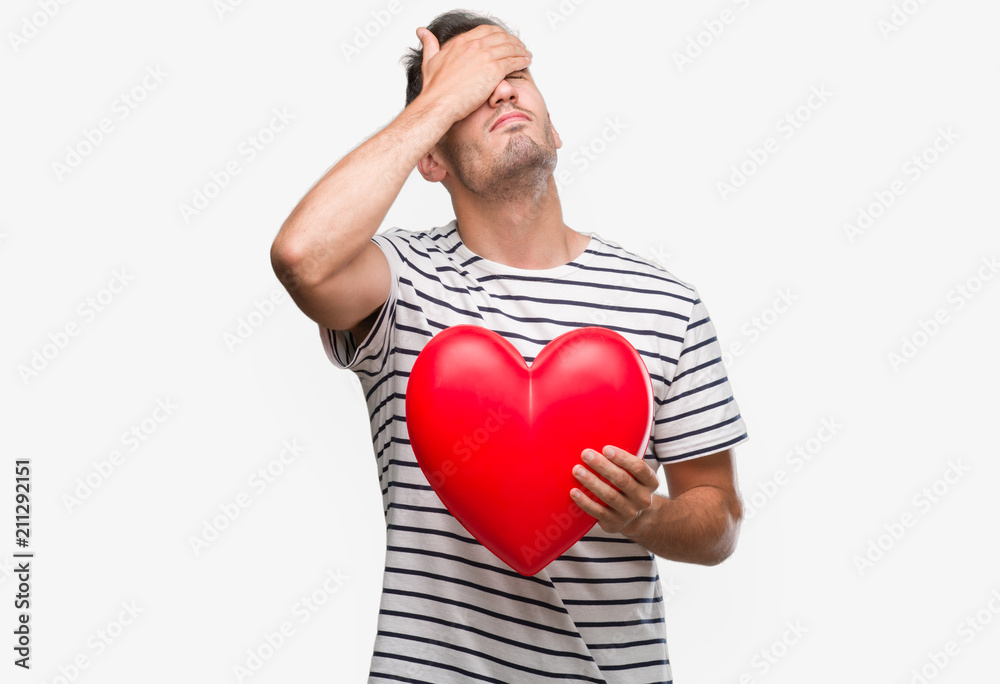 Handsome young man in love holding red heart stressed with hand on head, shocked with shame and surprise face, angry and frustrated. Fear and upset for mistake. Handsome young man in love holding red heart stressed with hand on head, shocked with shame and surprise face, angry and frustrated. Fear and upset for mistake.