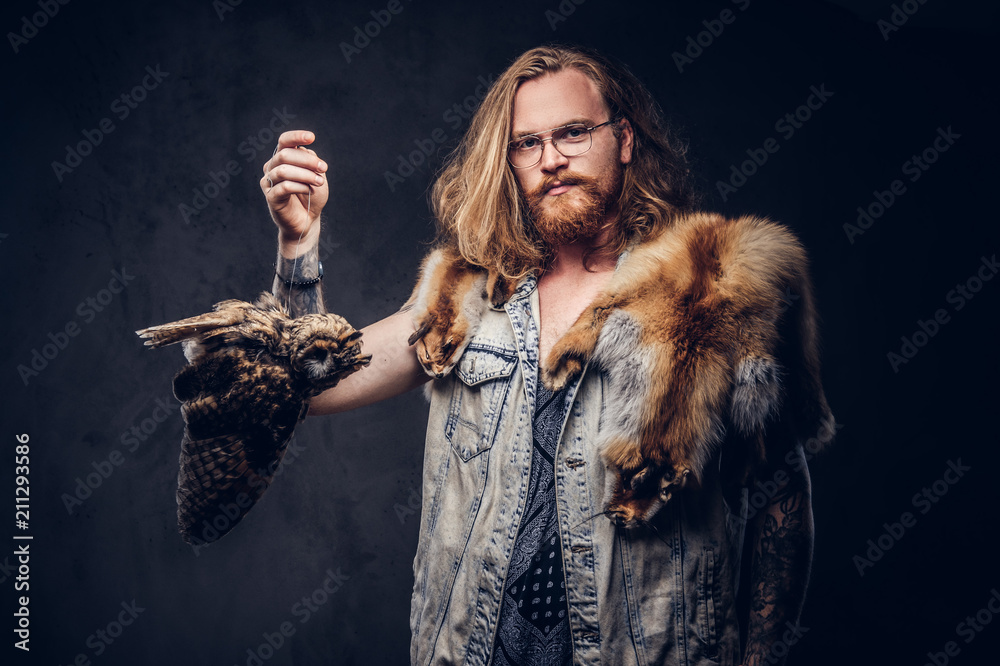 Portrait of a tattoed redhead hipster male with long luxuriant hair and full beard dressed in a t-shirt and jacket holds a keeps the scarecrow of an owl and fox skin on a shoulder in a studio.