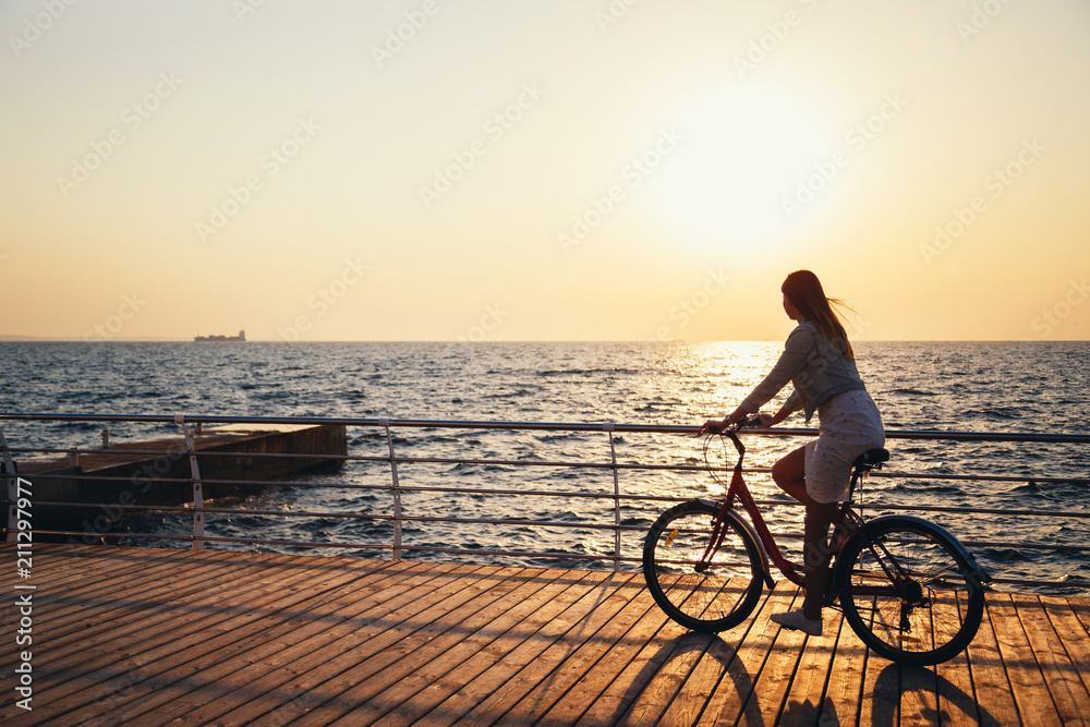 Young woman cycling at the beach at sunrise sky at wooden deck summer time