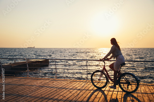 Young woman cycling at the beach at sunrise sky at wooden deck summer time