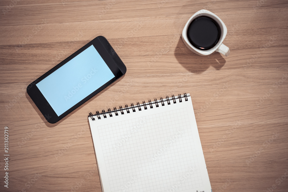 On the table is a smartphone with a blue screen and a laptop with a pen. There is coffee in the cup. On the table is a smartphone with a blue screen and a laptop with a pen. There is coffee in the cup.
