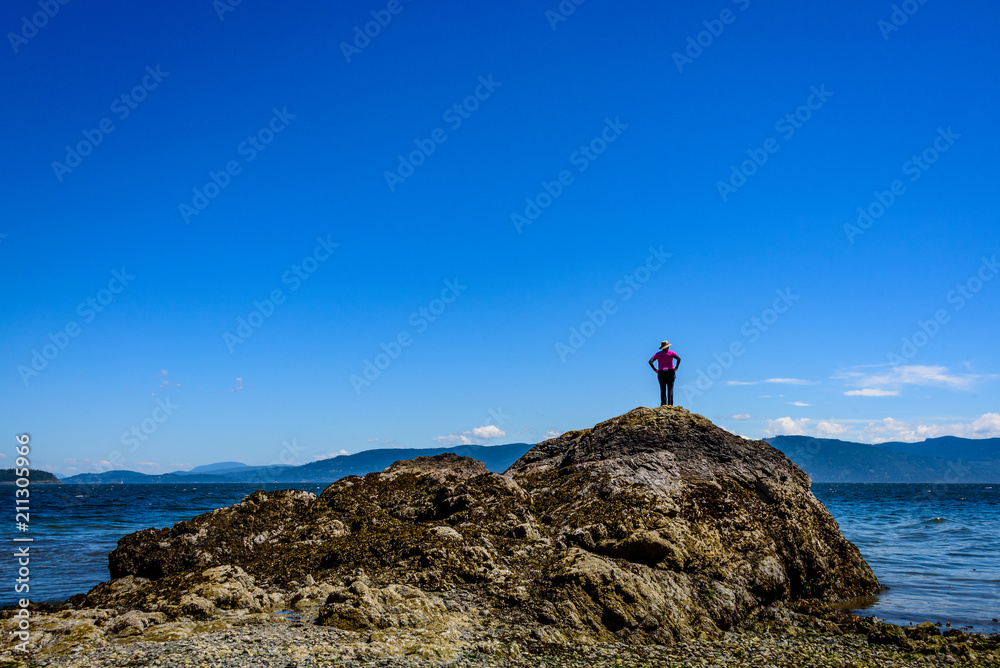 Woman in black pants and pink shirt climbing a rock formation on a beach in the San Juan Islands, the Salish Sea and other islands in the background, sunny day with a blue sky and while clouds
