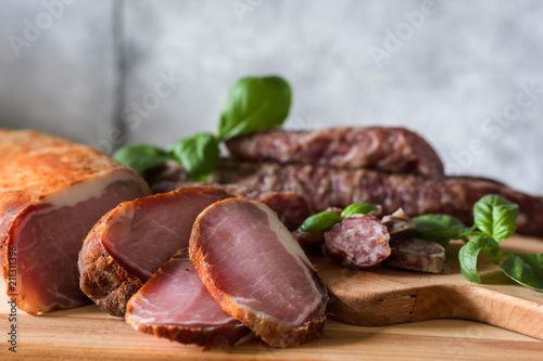 Dry home-made sausages and meat, serving slices on a wooden background decorated with a green basil on a gray background. Soft focus.