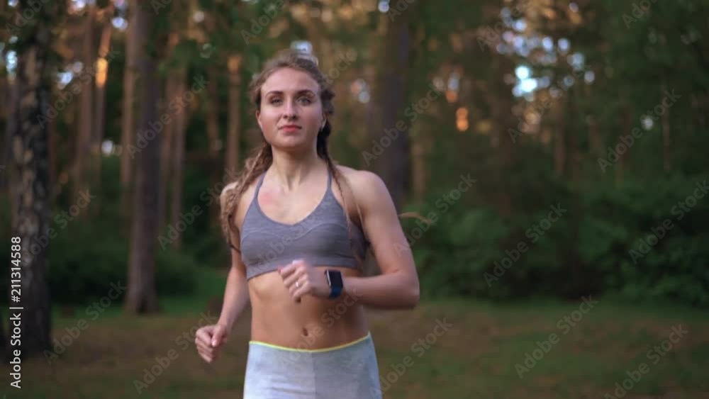 Front view of a slim caucasian woman in gray sportswear jogging in summer woods. Tracking slow motion medium shot Front view of a slim caucasian woman in gray sportswear jogging in summer woods. Tracking slow motion medium shot