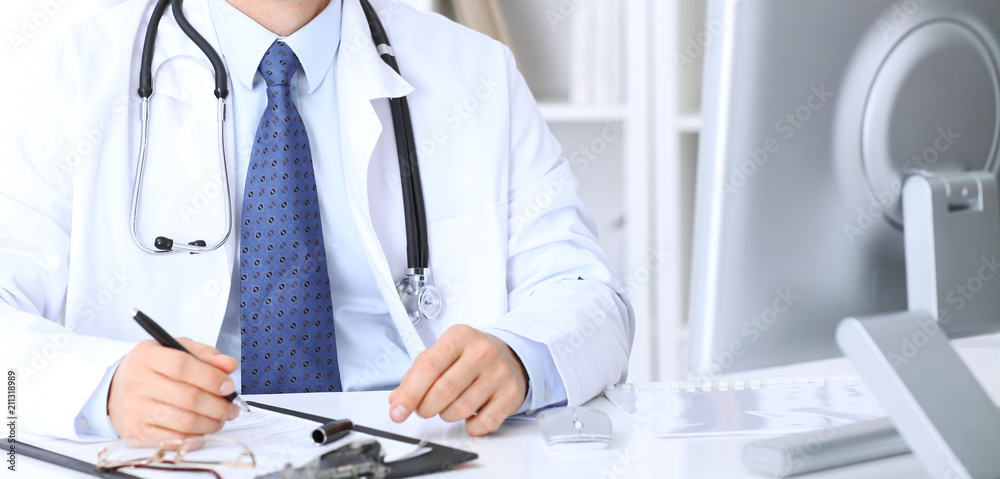 Unknown male doctor is sitting at the table and working in the hospital office. Closeup of stethoscope Unknown male doctor is sitting at the table and working in the hospital office. Closeup of stethoscope