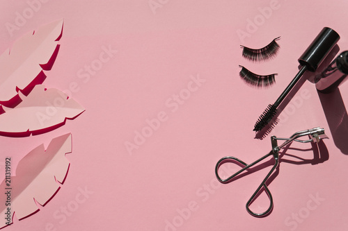 Black false lashes strips,mascara, curler on pink background with paper leaves photo