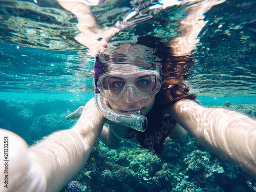 Young woman taking an underwater selfie wearing snorkeling mask when swiming and diving in red sea with clear turquoise blue water. Coral reef