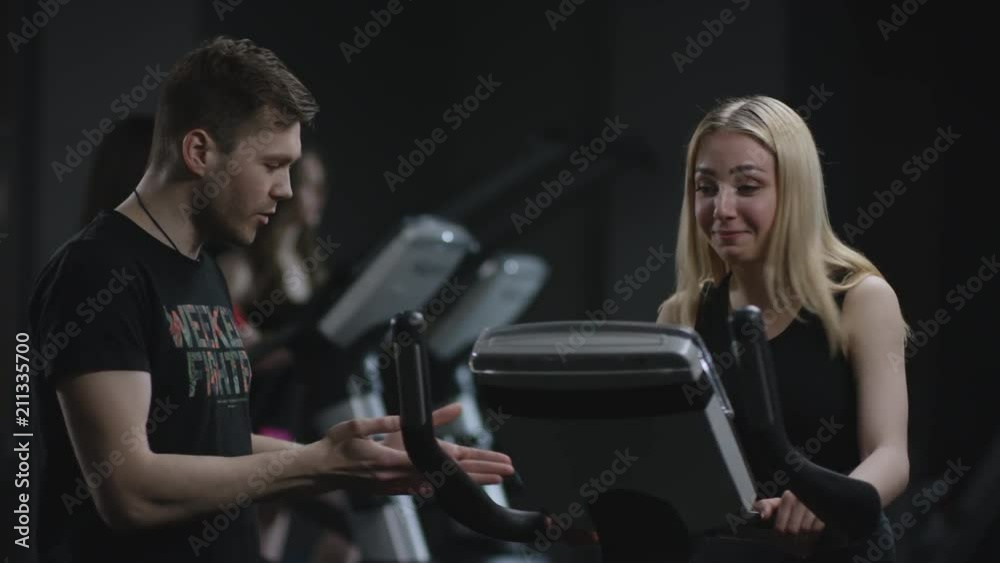 nice blonde girl is diligent working on an exercise bike and cool guy come to her and suggests to make selfie on a background of practiced girls
