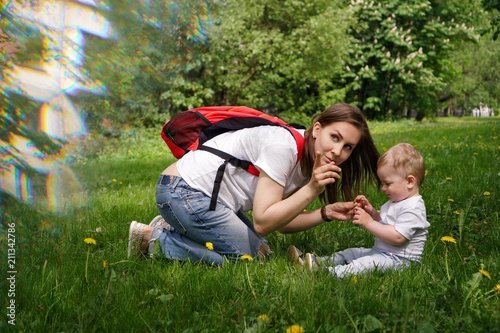 Family. Son and mother outdoors. Pregnant girl with backpack is sitting on lawn and showing to side. Little boy is playing. Blur effect