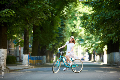 Positive pretty girl in white dress and straw hat is happy riding blue bike down wide beautiful park alley with trees around on sunny summer day. Beautiful female ride enjoying happiness healthy © anatoliy_gleb