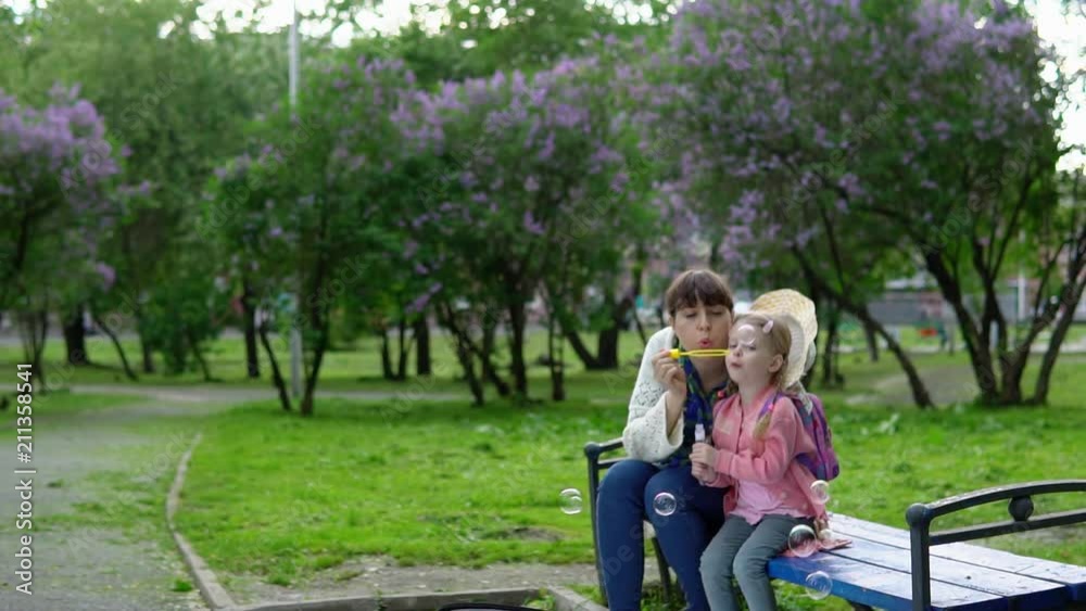 Young mother and cute daughter blowing soap bubbles in park of blooming lilacs on sunny spring day, slow motion. Young mother and cute daughter blowing soap bubbles in park of blooming lilacs on sunny spring day, slow motion.