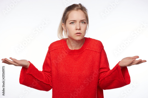 Portrait of puzzled intelligent female student wears red oversized sweater, feels awkward, shrugs shoulders with hesitation, sees no way out, not knowing material on exam isolated on white wall.