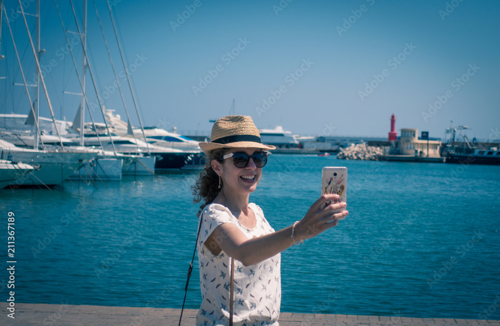 Happy young long-haired tourist woman blogger wearing black hat and round sunglasses is smiling while making a selfie with a cell phone