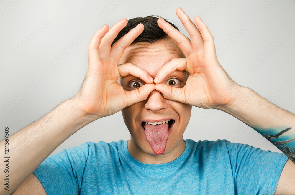 Young guy dressed in a blue t-shirt looks through binoculars from the palms of his hands and shows tongue, isolated on a light background.