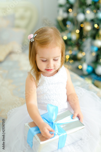 Little girl sitting on bed with present and wearing white dress, decorated fir tree in background. Concept of winter holidays and New Year gifts. photo