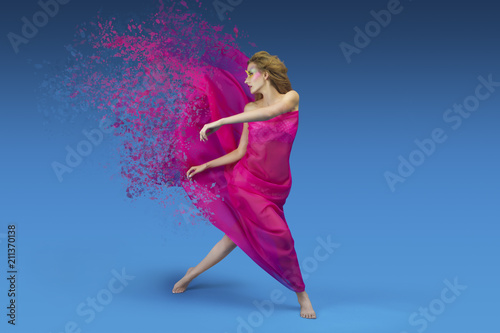 Fashionable girl with ginger hair and bright make up posing / dancing with pink and silky fabric on blue background. Dispersion effect photo