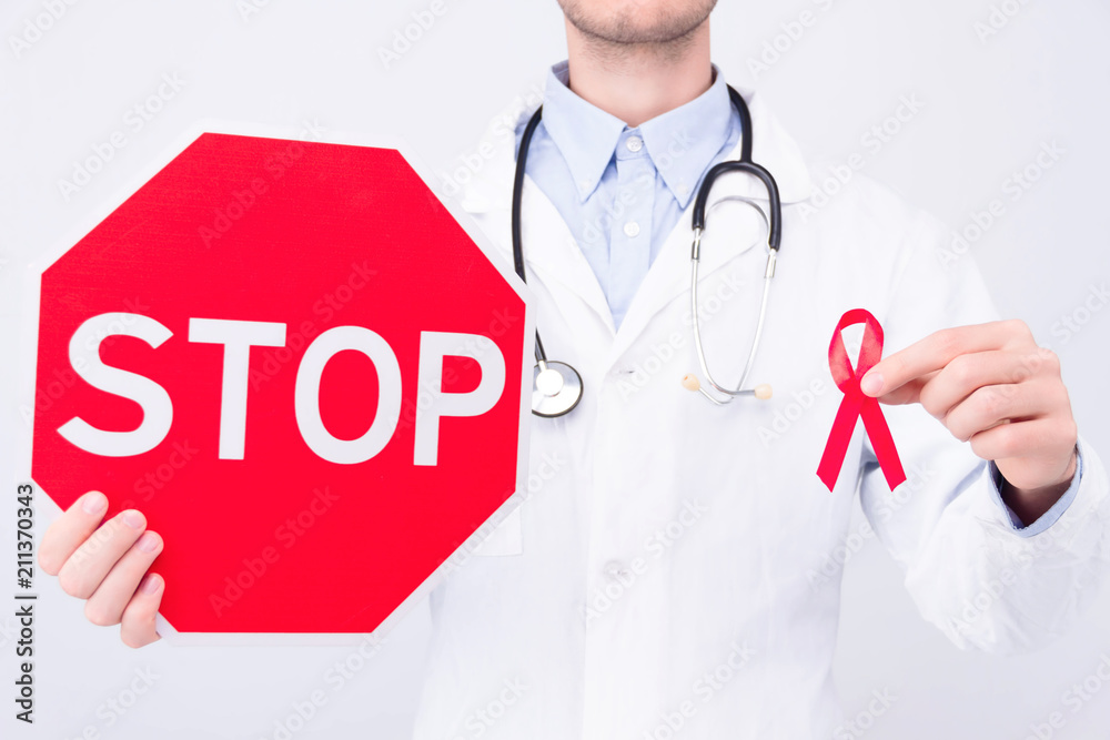 Oncological disease concept. Doctor wearing white coat and stethoscope holding red ribbon as a symbol of aids and stop sign Oncological disease concept. Doctor wearing white coat and stethoscope holding red ribbon as a symbol of aids and stop sign