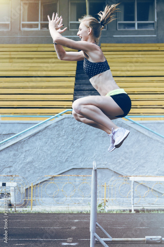 Fototapeta Naklejka Na Ścianę i Meble -  Athletic girl wearing sneakers and sport suit jumping above the hurdle on stadium running track