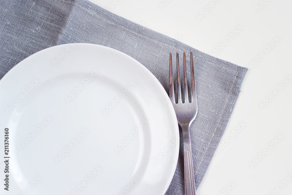 Empty white plate on a gray napkin with fork on white table. Table setting, preparation for food.