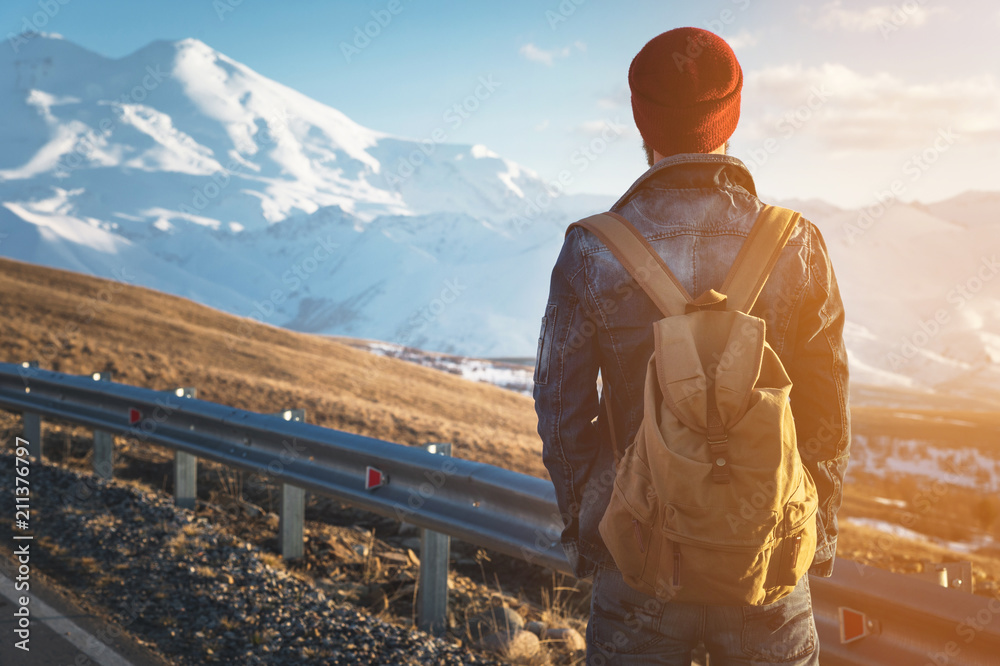 Bearded tourist hipster man in sunglasses with a backpack stand back on a roadside bump and watching the sunset against the background of a snow capped mountain - obrazy, fototapety, plakaty 