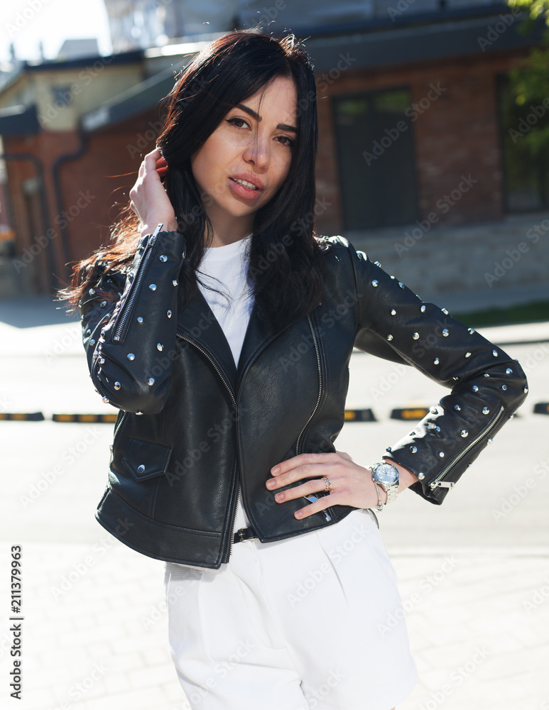 Fashionable brunette girl blogger posing in a black jacket, white t-shirt and white shorts on a sunny street in the evening. Content.