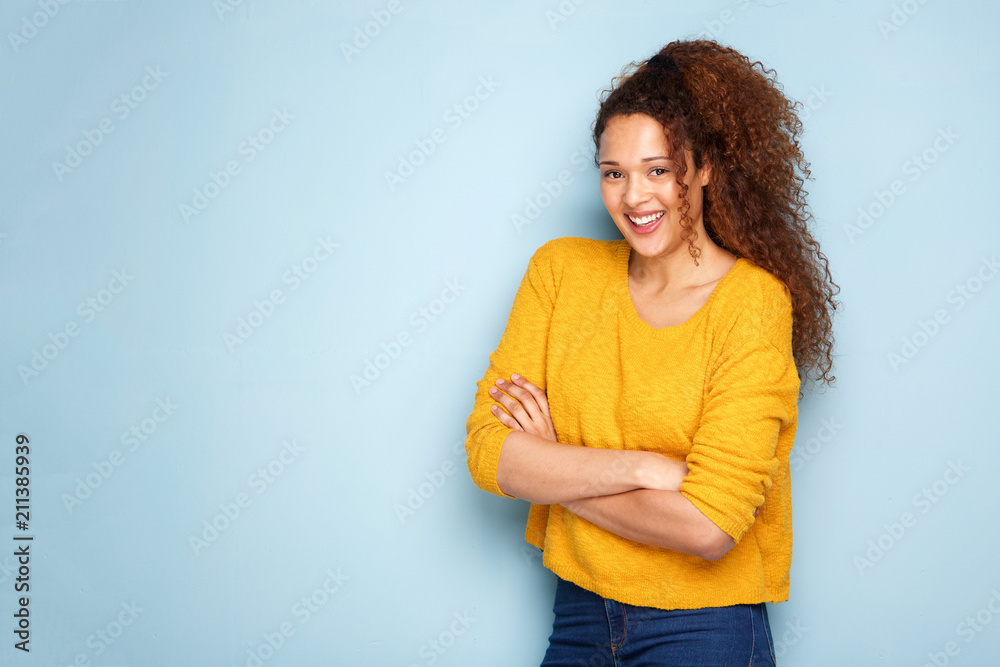 young woman with curly hair smiling by blue wall with arms crossed young woman with curly hair smiling by blue wall with arms crossed