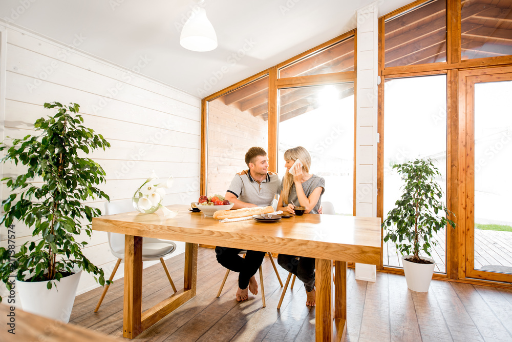 Young and happy couple having a breakfast sitting at the wooden table in the modern country house with big window on the background Young and happy couple having a breakfast sitting at the wooden table in the modern country house with big window on the background