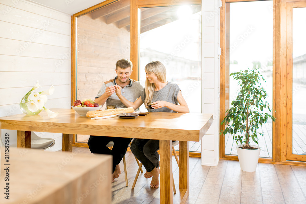 Young and happy couple having a breakfast sitting at the wooden table in the modern country house with big window on the background Young and happy couple having a breakfast sitting at the wooden table in the modern country house with big window on the background