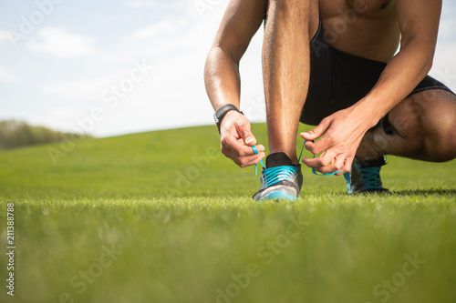 Close-up of shapely guy is stringing sneakers laces. Shredded guy is spending warm sunny day in green environment. Man is using smartwatch