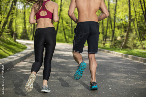 Athletic man and woman are running on alley among green environment. They are having cardio training jointly on lovely warm day. Male and female are turning back to camera