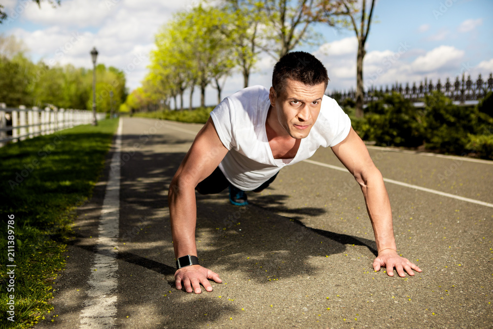 Athletic male with tracker is working out on alley in park. He is doing press-ups while feeling very concentrated. Power training outside concept Athletic male with tracker is working out on alley in park. He is doing press-ups while feeling very concentrated. Power training outside concept