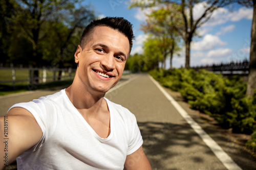 Portrait of cheerful handsome guy smiling to camera while making selfie. He is enjoying free time while walking along asphalt road in sunny park