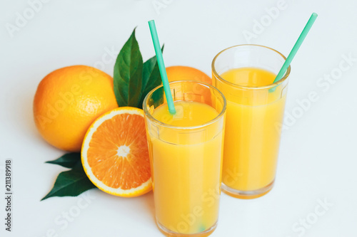 Glasses with orange juice next to orange slices with leaves on a white background.
