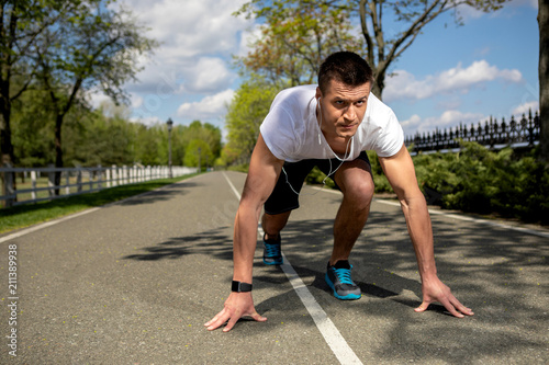 Athletic man is bending to asphalt in crouch start. He is getting ready for running along sunny road in park while feeling very concentrated. Male is wearing smartwatch and using headphones