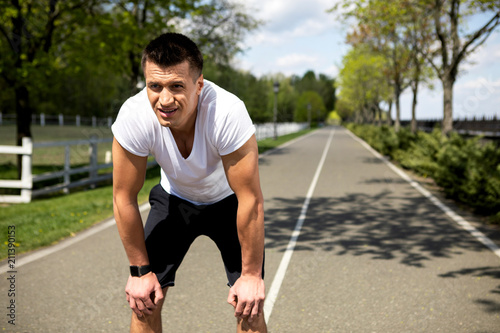 Strong man with tracker is knackered after cardio exercises on track among green trees. He is bending for short relaxation while standing on sunny road and looking ahead. Copy space in right side